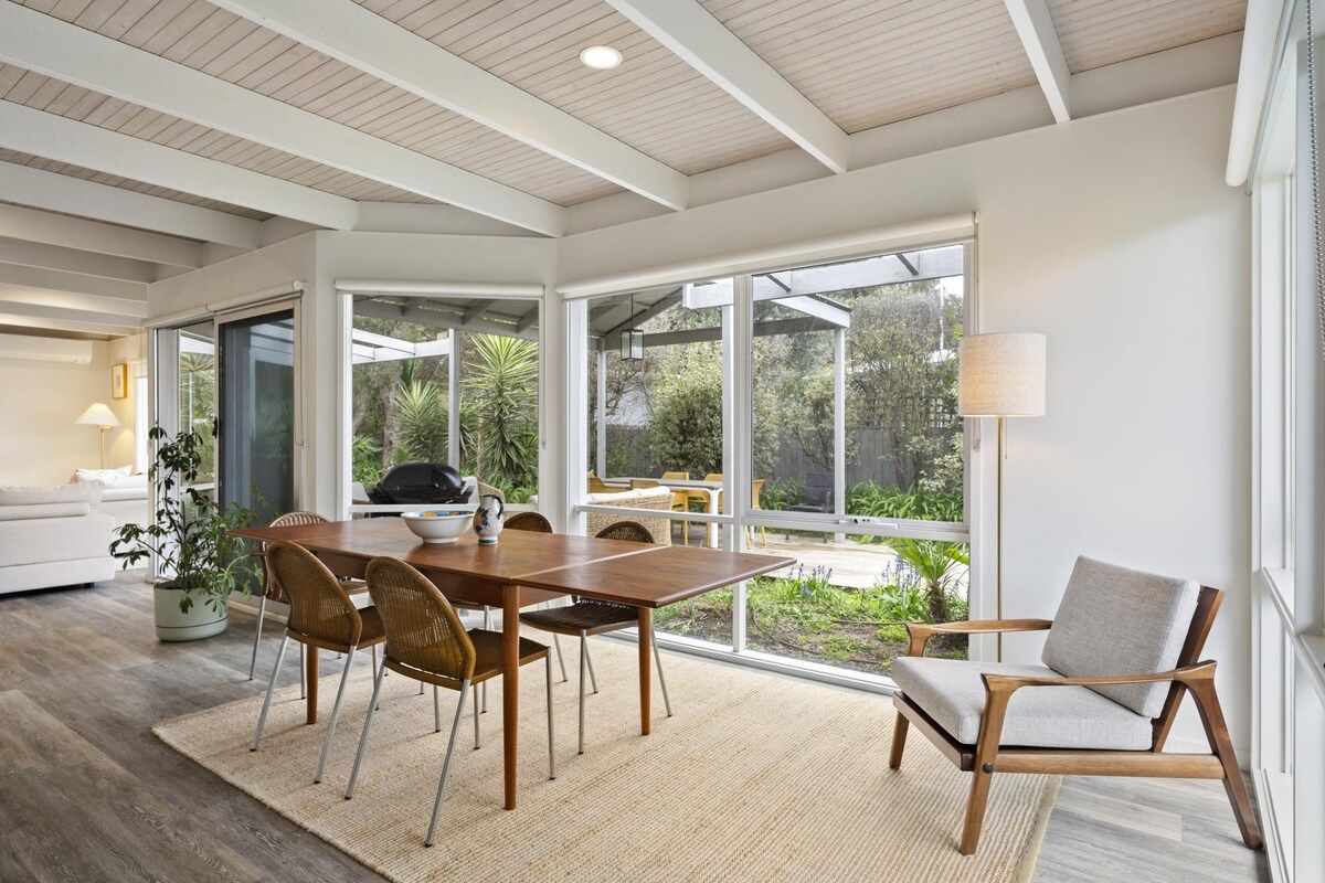 Un comedor moderno con una mesa y sillas de madera, ventanas amplias que ofrecen vista a follaje, y una lámpara a la derecha.
