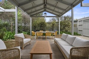 Een terras buiten met rieten meubels, een houten tafel en een eethoek met gele stoelen.