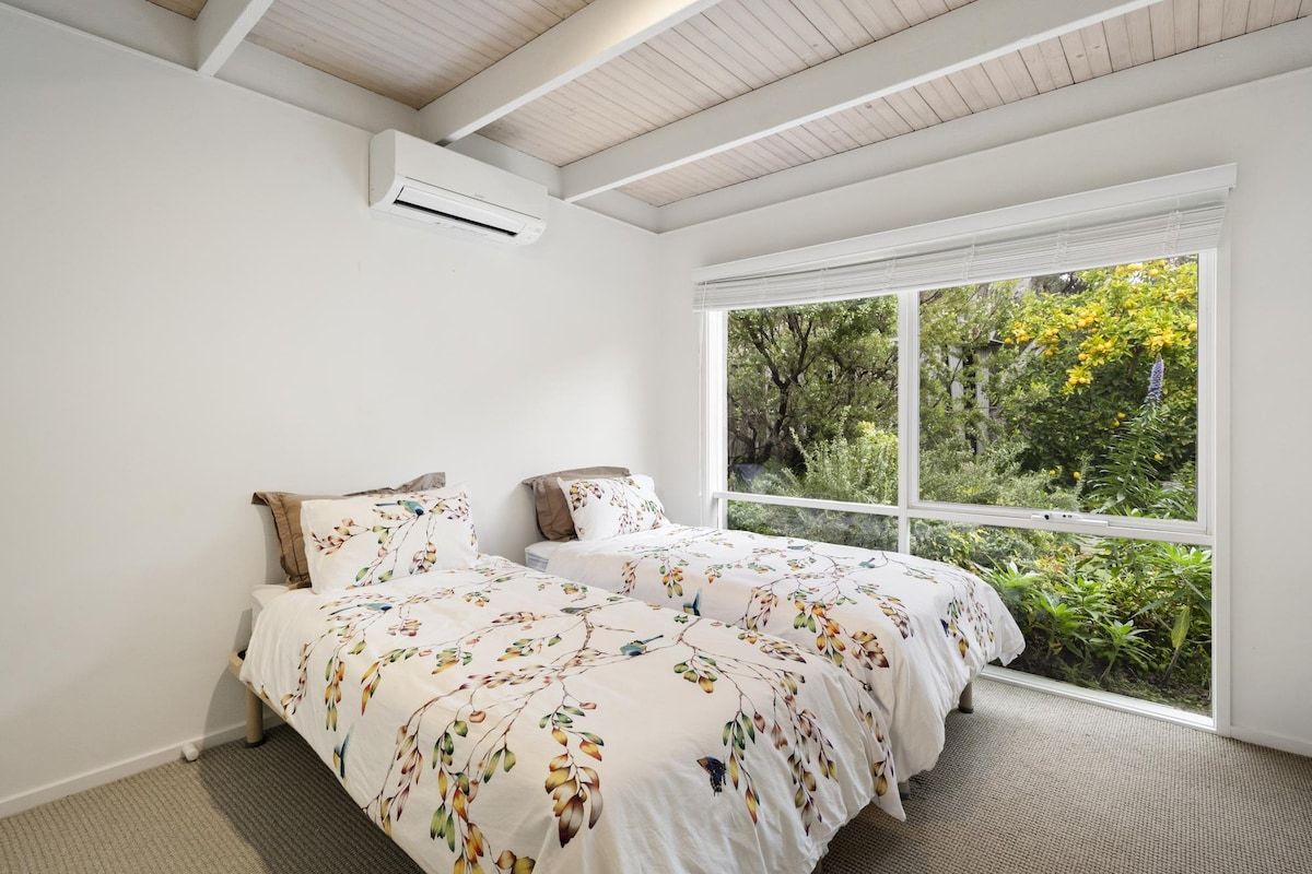 Une chambre avec une grande fenêtre, un lit recouvert d’une literie à motifs floraux et un climatiseur fixé au mur.