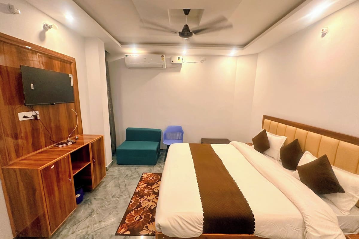A hotel room with a bed, a TV mounted on a wooden cabinet, a ceiling fan, and a small seating area with a blue chair.