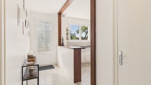 Un couloir avec une porte en bois, une porte en verre et une fenêtre donnant sur des arbres. On y trouve une petite table sur laquelle sont posés un panier et une boîte.