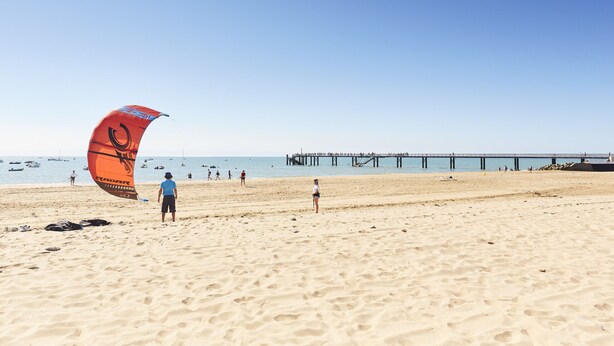 Une plage avec une jetée en arrière-plan et une personne faisant voler un cerf-volant.