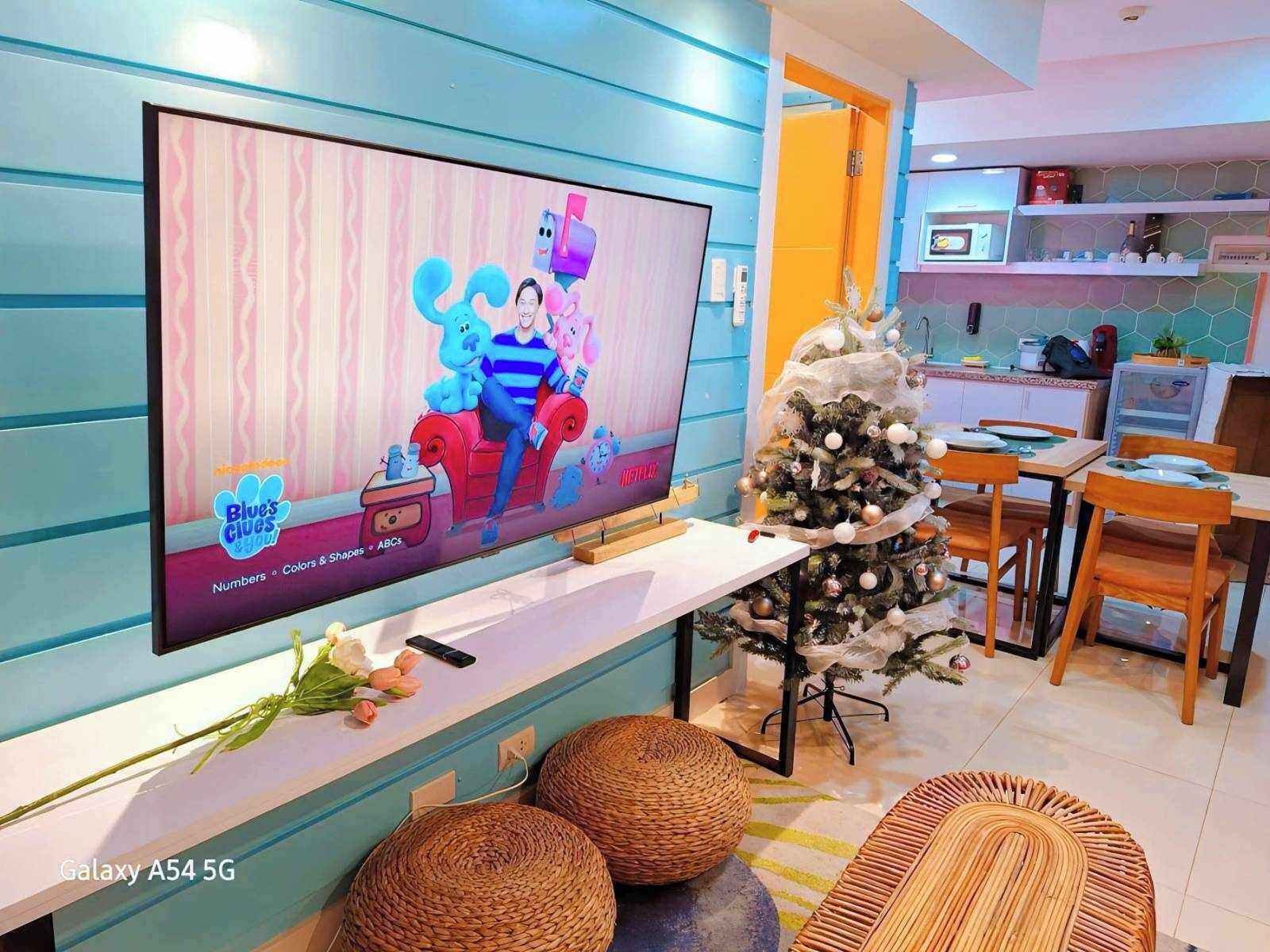 A living room with a flat-screen TV, a Christmas tree, wicker ottomans, and a kitchen area in the background.