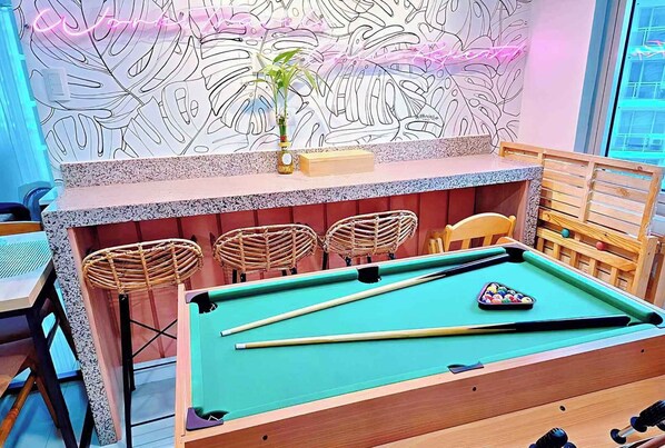 A pool table with a neon sign, a plant, and wicker chairs.