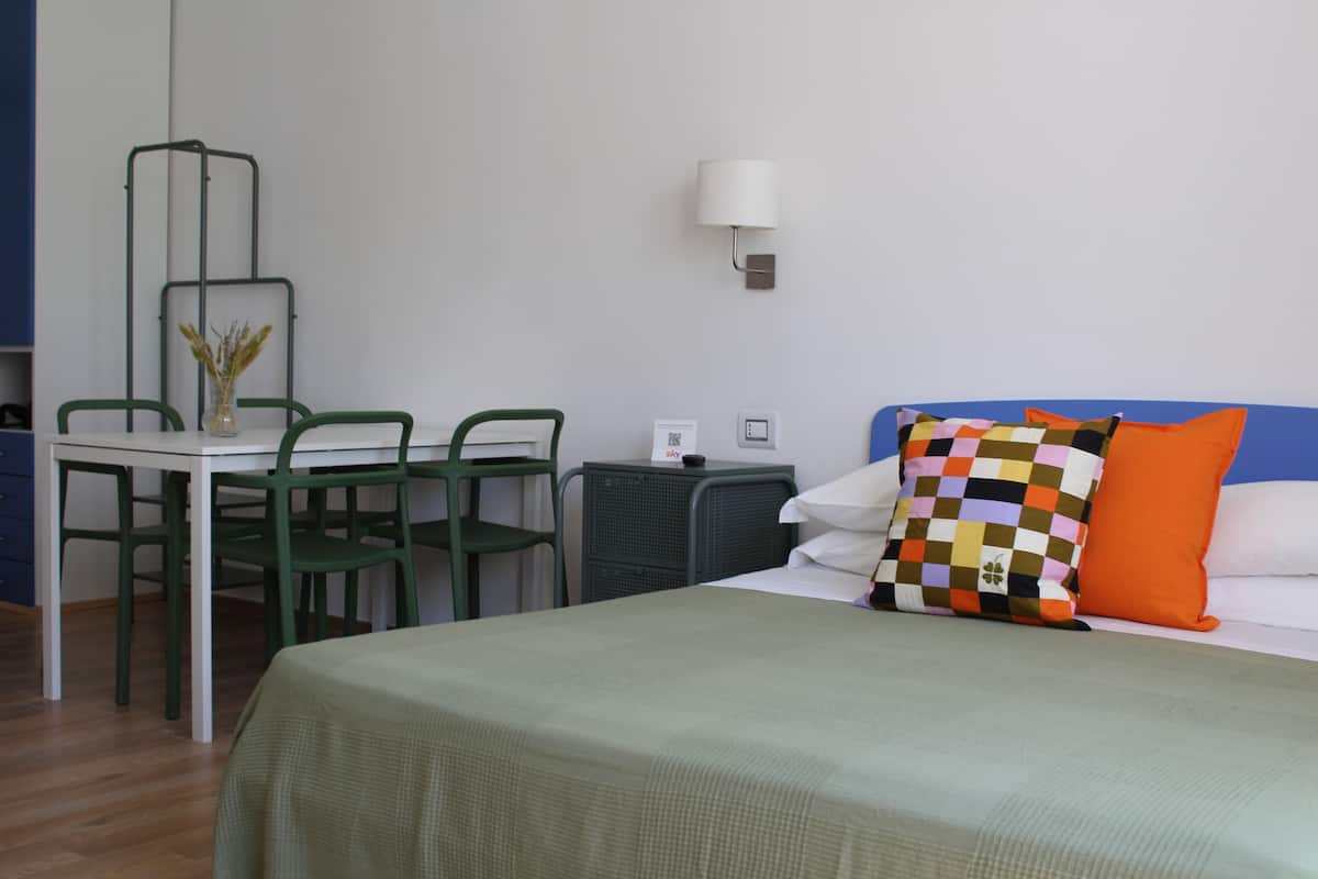 A hotel room with a bed, a table with chairs, a lamp, and a small desk with a computer.