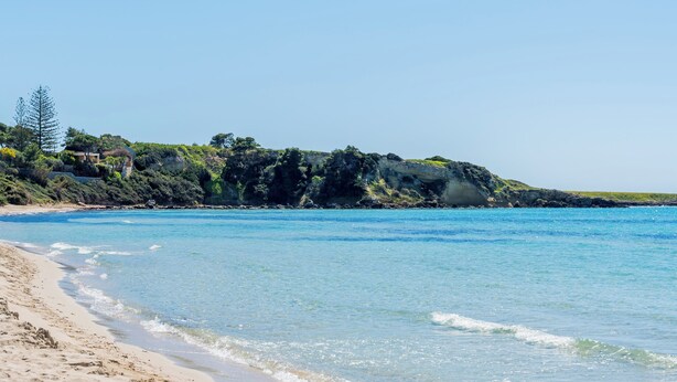 Ein Sandstrand mit klarem, blauem Wasser und einem Felsen im Hintergrund.