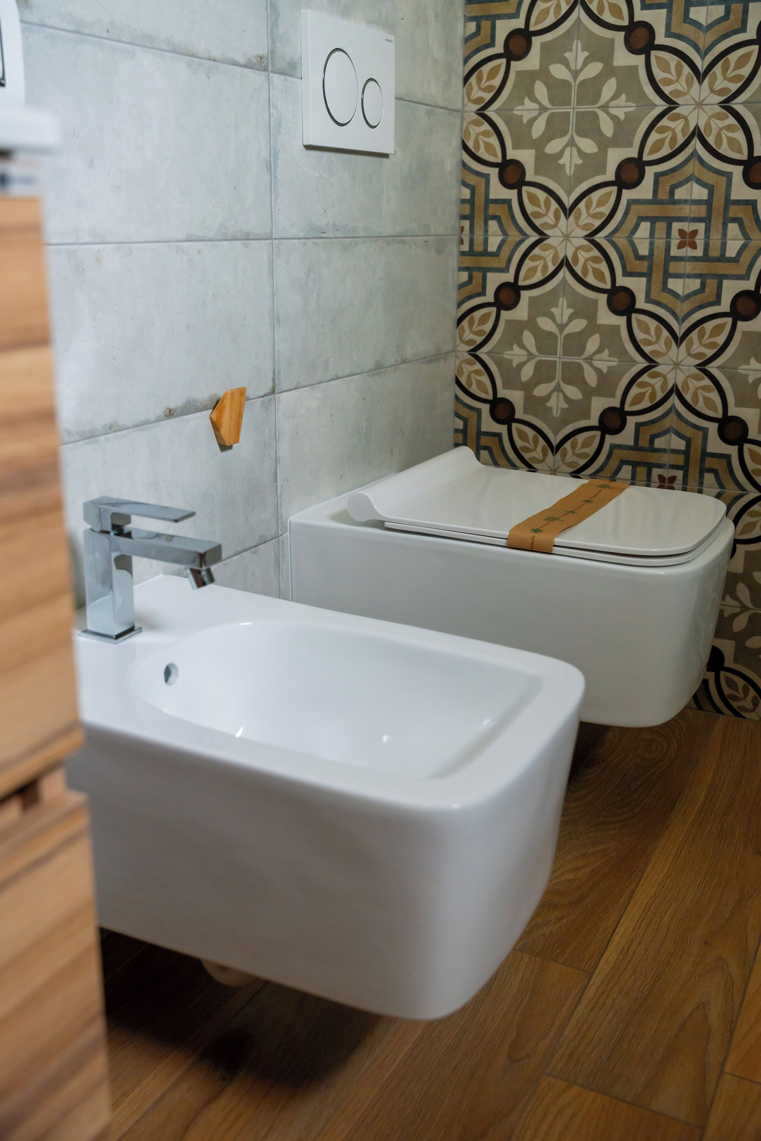 A bathroom with a white sink, a toilet, and a bidet. The walls are tiled, and there is a patterned wallpaper on one side.
