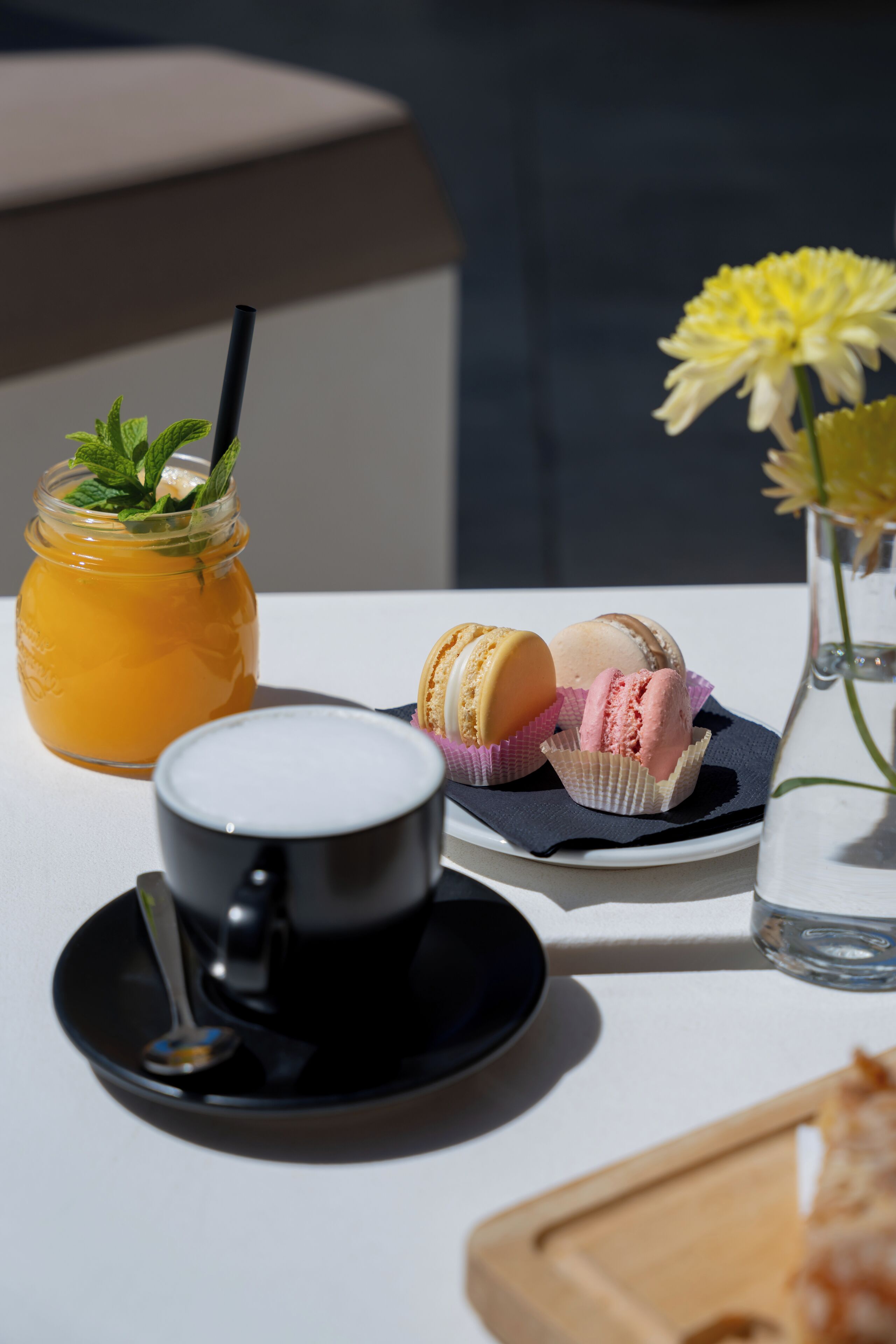 Une table avec une tasse noire, un verre de boisson orange, des macarons et un vase rempli de fleurs.