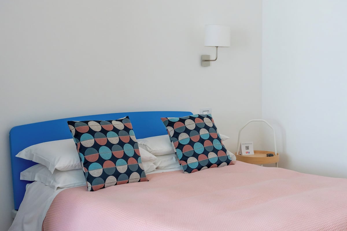 A neatly made bed with a blue headboard, white and patterned pillows, and a pink bedspread. A small bedside table with a lamp and a charging station is visible.