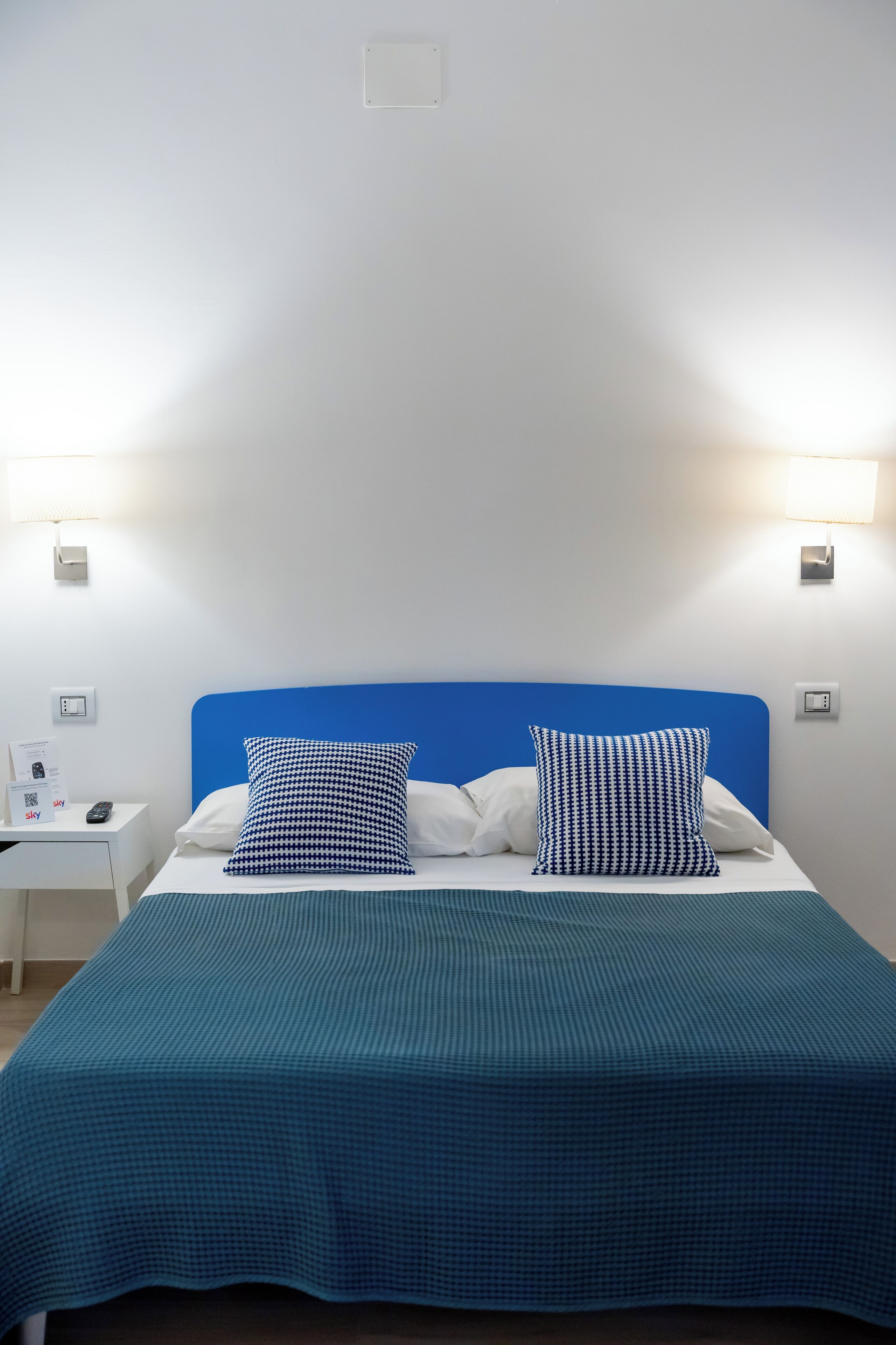 A hotel room with a bed, two pillows, a nightstand, and a wall-mounted lamp.