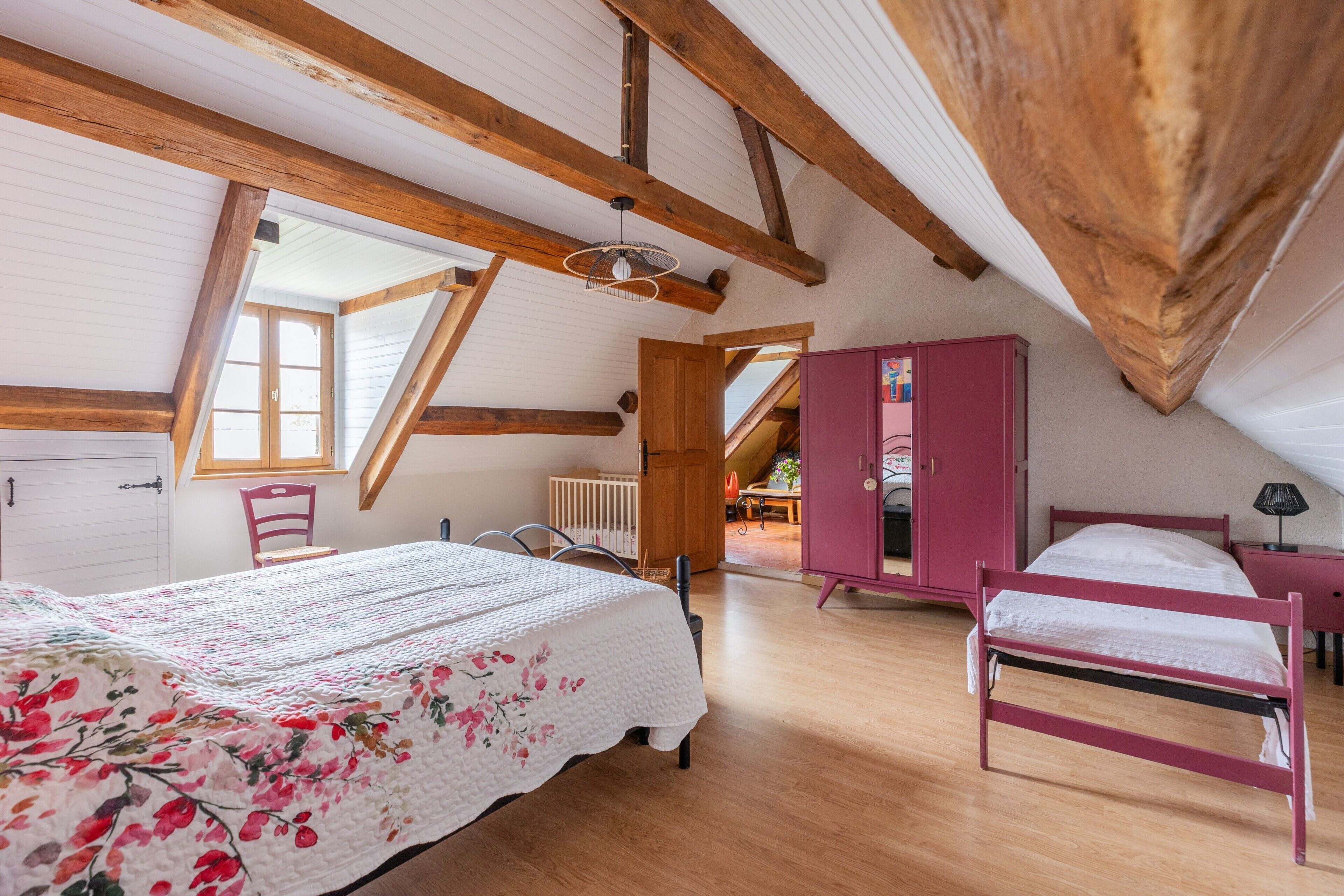 Et soveværelse med træloft, en seng med blomstret dynebetræk, en rosa garderobe og et vindue.