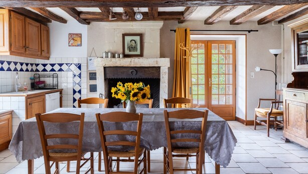 Une cuisine rustique avec un mur recouvert de carreaux, des armoires en bois, une table à manger avec des chaises, une cheminée décorée de tournesols et une fenêtre avec des rideaux.