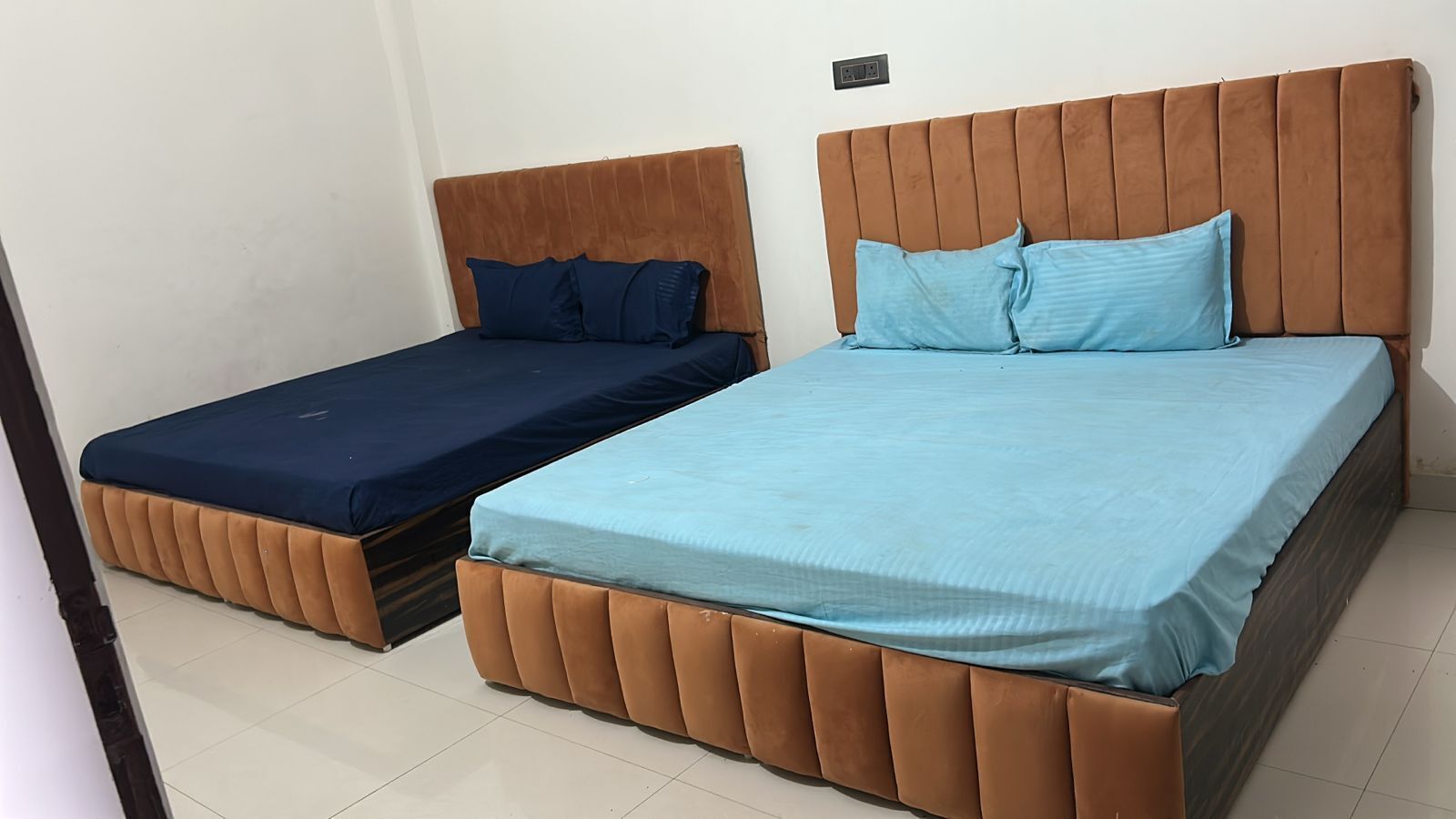 Ein Zimmer mit zwei Betten, einem größeren und einem kleineren, jeweils mit braunen Kopfteilen und blauer Bettwäsche.