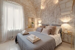 A bedroom with a stone wall, a bed with a grey quilt, pillows, a bedside table with a lamp, and a window with sheer curtains.