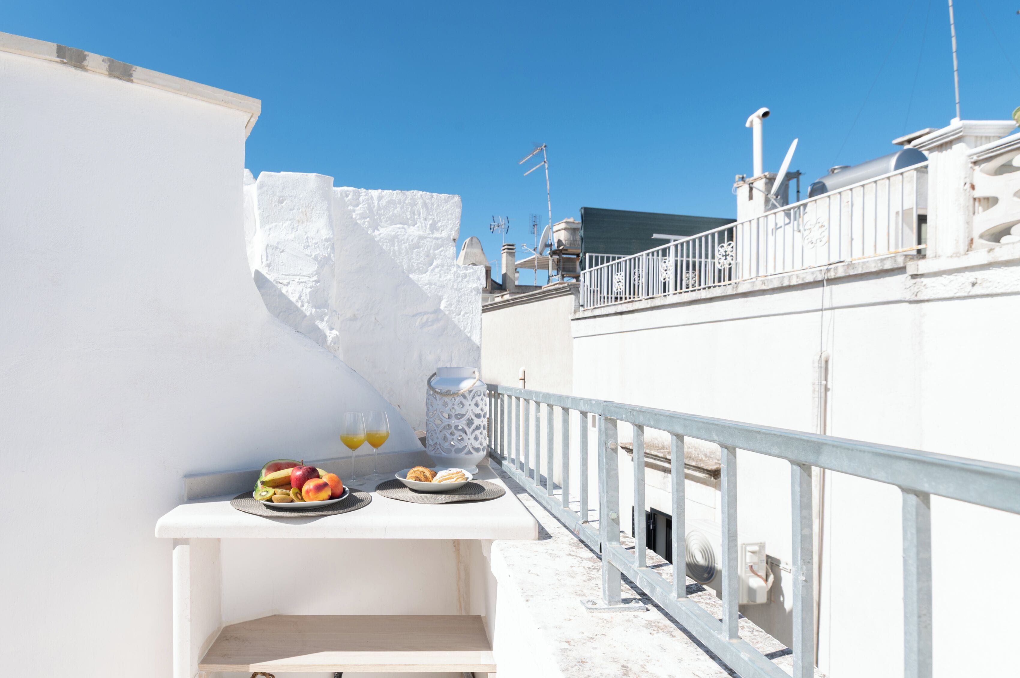 Una terrazza sul tetto con un tavolo bianco apparecchiato con frutta, bicchieri di succo e un vaso decorativo.