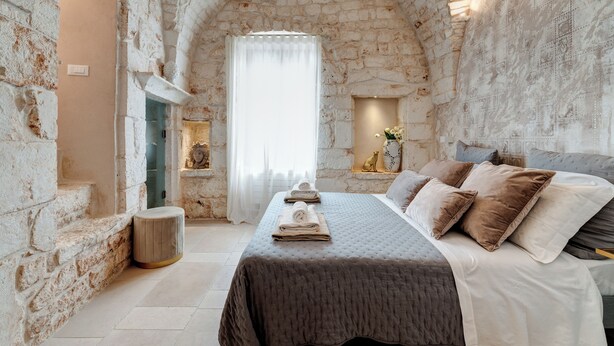 A stone-walled room with a bed, a small stool, and a window with curtains.