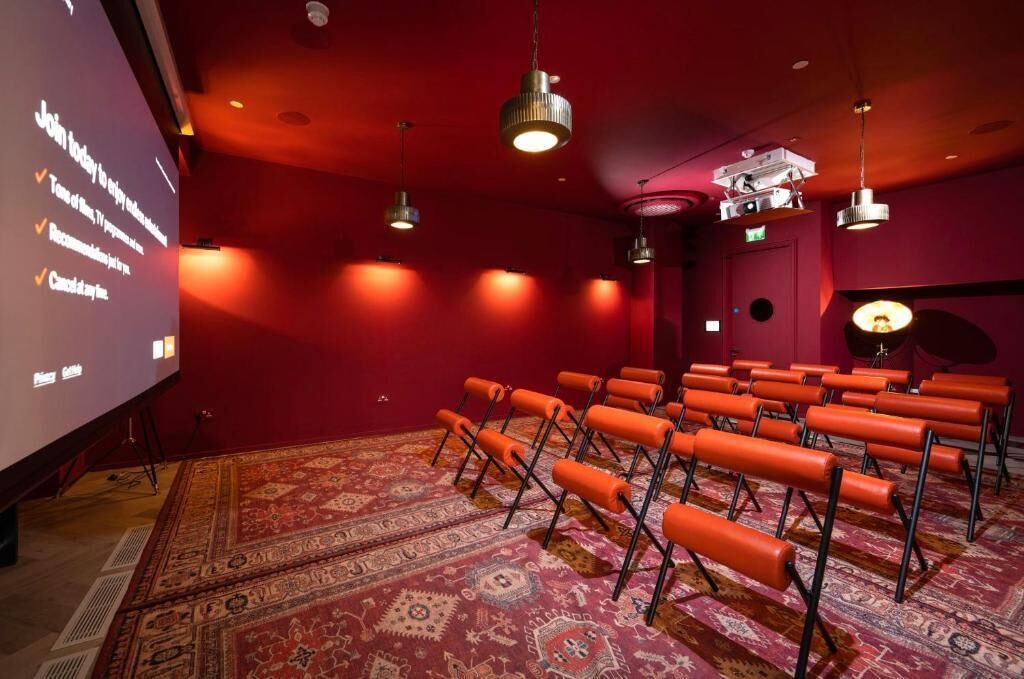Une salle de théâtre avec des rangées de sièges orange, un grand écran et des murs rouges.