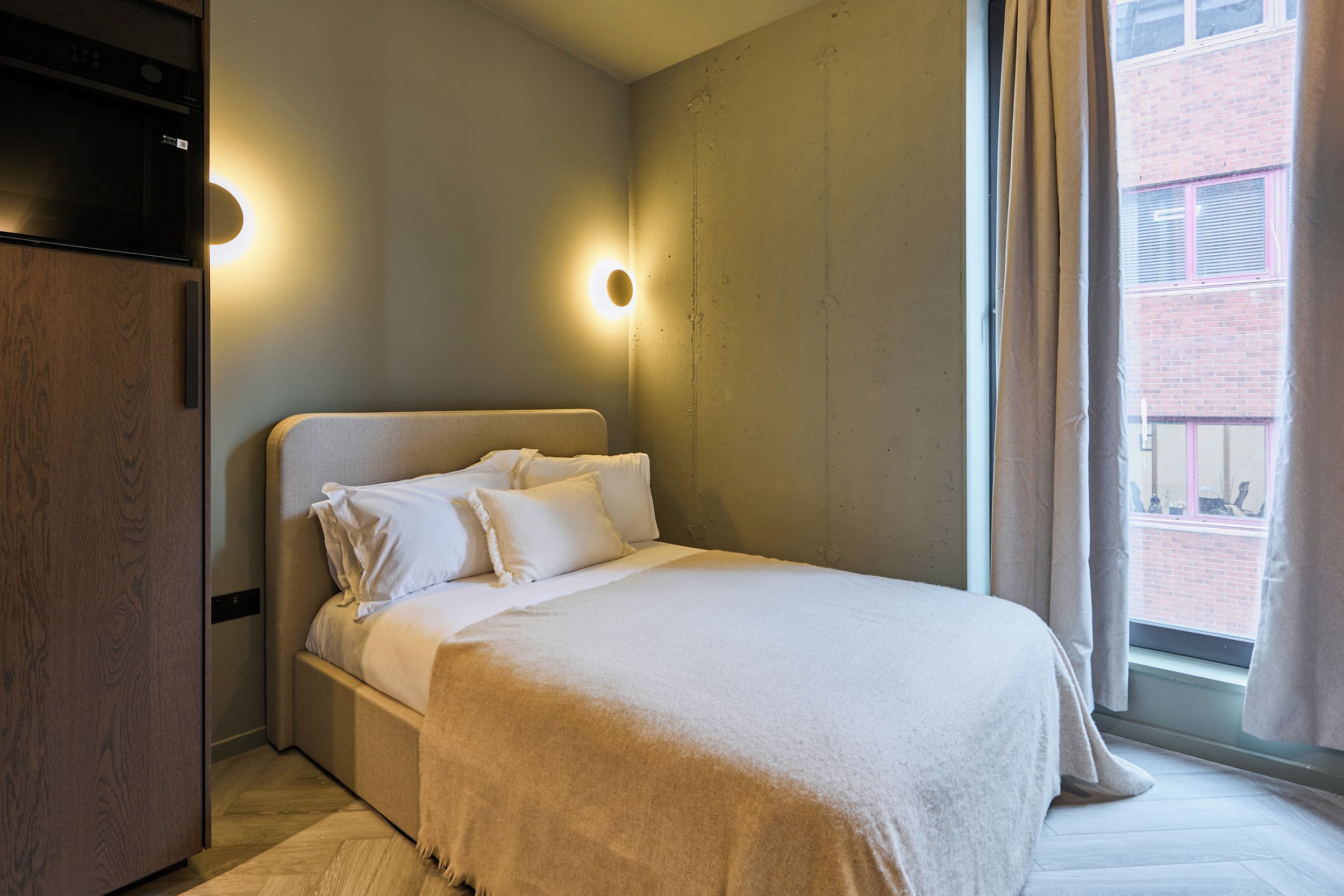 Une chambre d’hôtel avec un lit, une armoire en bois, une lampe fixée au mur et une fenêtre avec des rideaux.