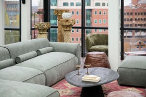 Una sala de estar moderna con un sofá gris, una mesa de centro redonda con un libro y dos vasos, y vistas a edificios de la ciudad a través de grandes ventanales.