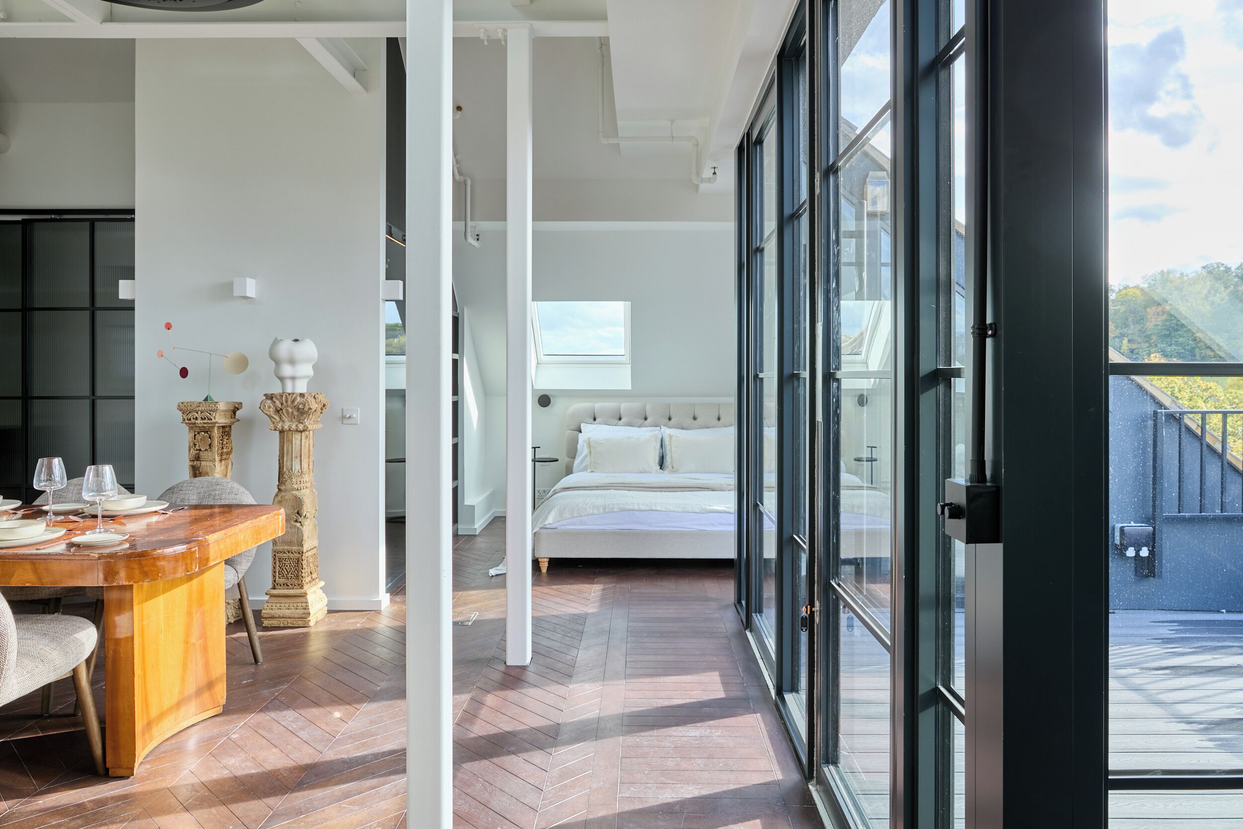 A modern bedroom with a large bed, a dining area with a wooden table, and a view through large windows.