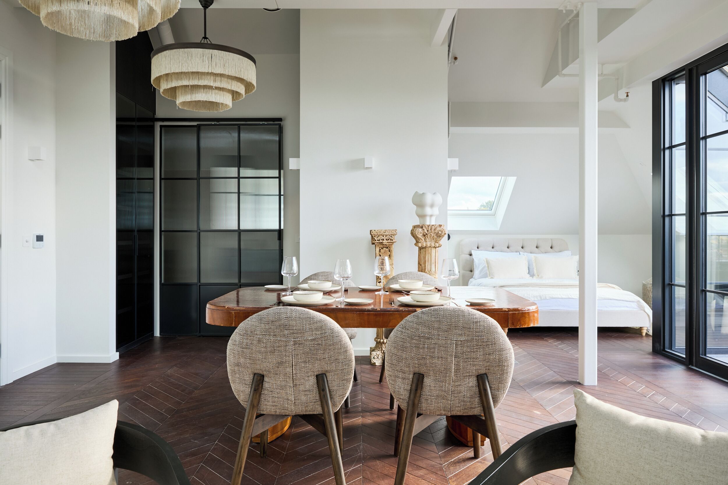 Un'area pranzo moderna con un tavolo in legno apparecchiato per quattro persone, circondato da sedie imbottite. Un'ampia finestra offre luce naturale, e si intravede un letto con biancheria bianca sullo sfondo.