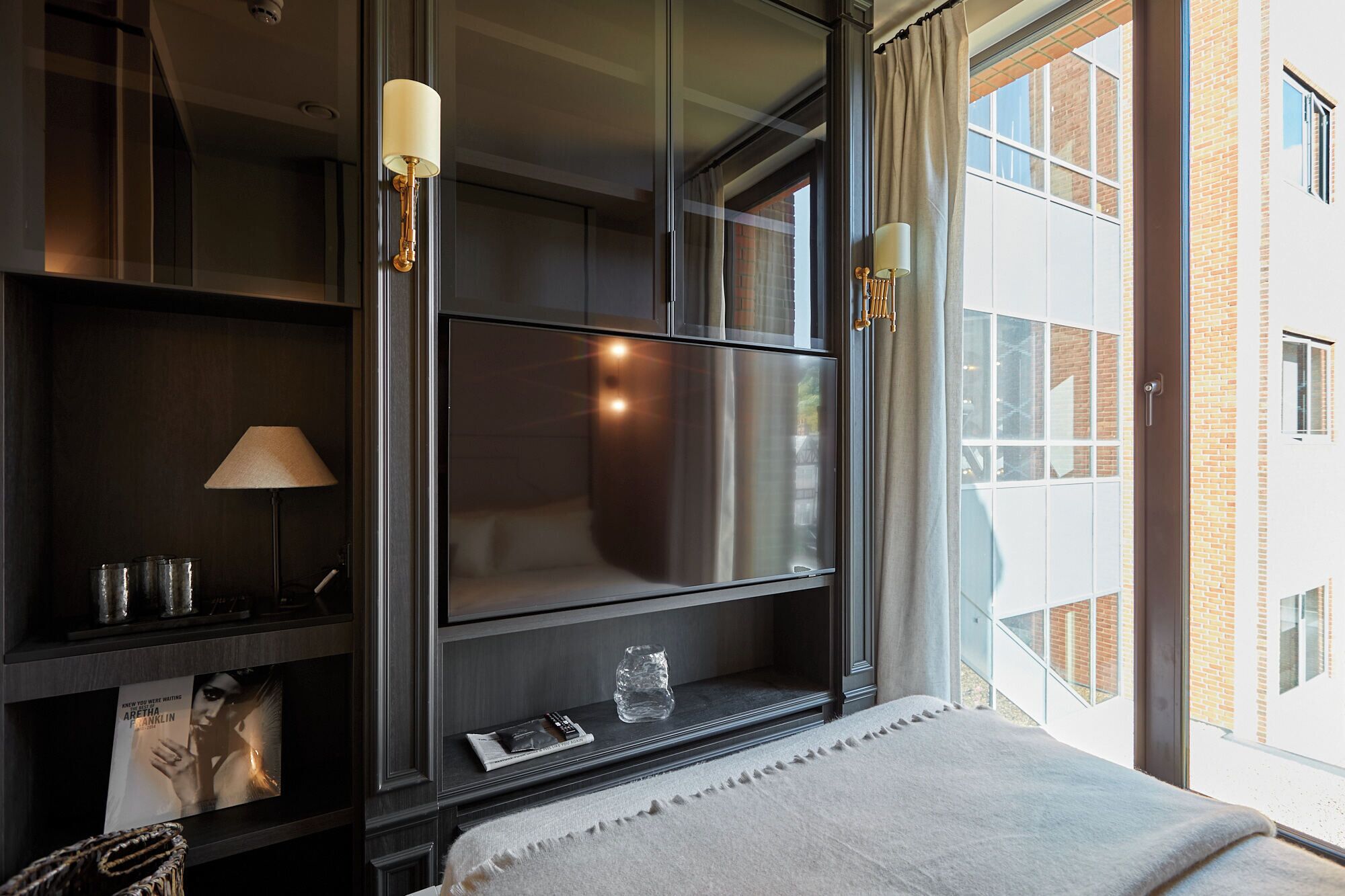Habitación de hotel moderna con un ventanal grande, un televisor de pantalla plana, una mesita de noche con lámpara y vistas a un edificio de ladrillo.