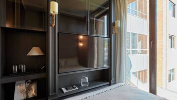 Habitación de hotel moderna con un ventanal grande, un televisor de pantalla plana, una mesita de noche con lámpara y vistas a un edificio de ladrillo.