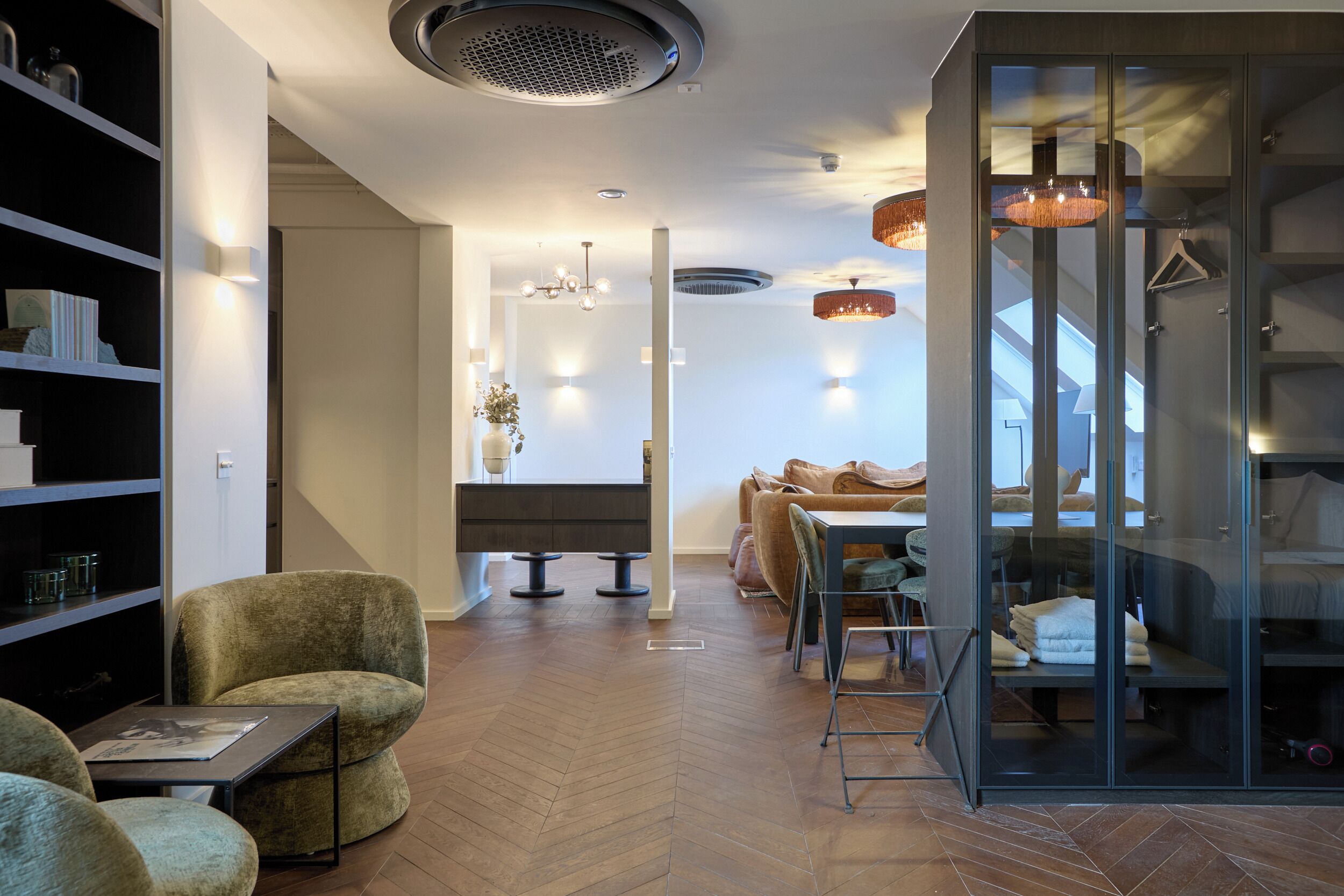 A modern interior with a herringbone wood floor, a sofa, a dining area, and a glass partitioned room with a bed.