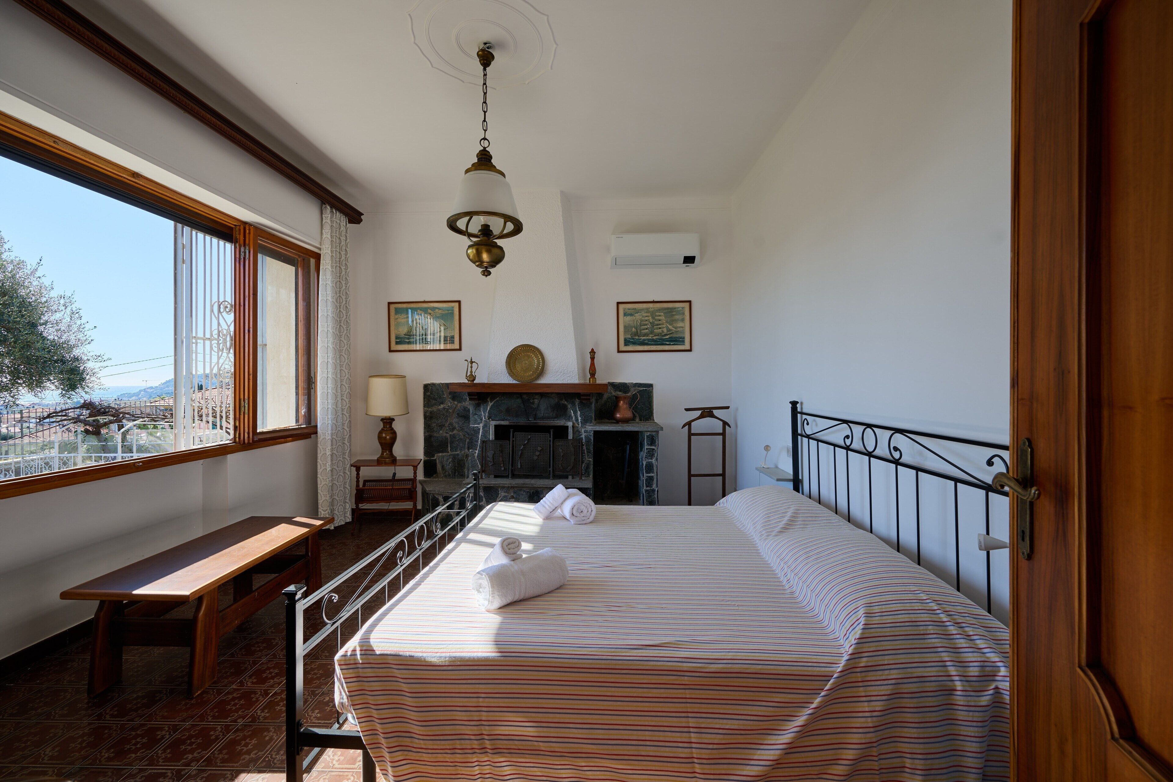 A bedroom with a bed, a bench, a fireplace, and a view through a window.