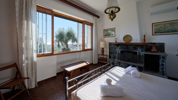 A bedroom with a large window, a wooden bench, a bed with a striped bedspread, a stone fireplace, and a hanging light fixture. - Villa Camilla App Garden (Diano Castello)
