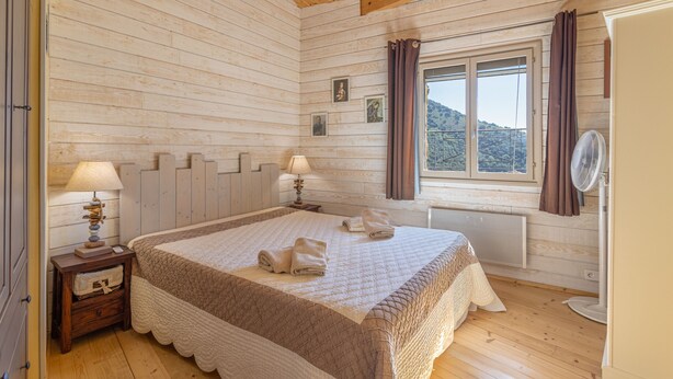 A wooden-paneled room with a bed, two bedside tables with lamps, a window with curtains, and a radiator. - Gîte Madres Coronat Balcon du Canigou (Jujols)