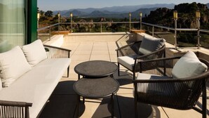 Una terrazza con divano, due tavolini e sedie in vimini, affacciata su un paesaggio montuoso.