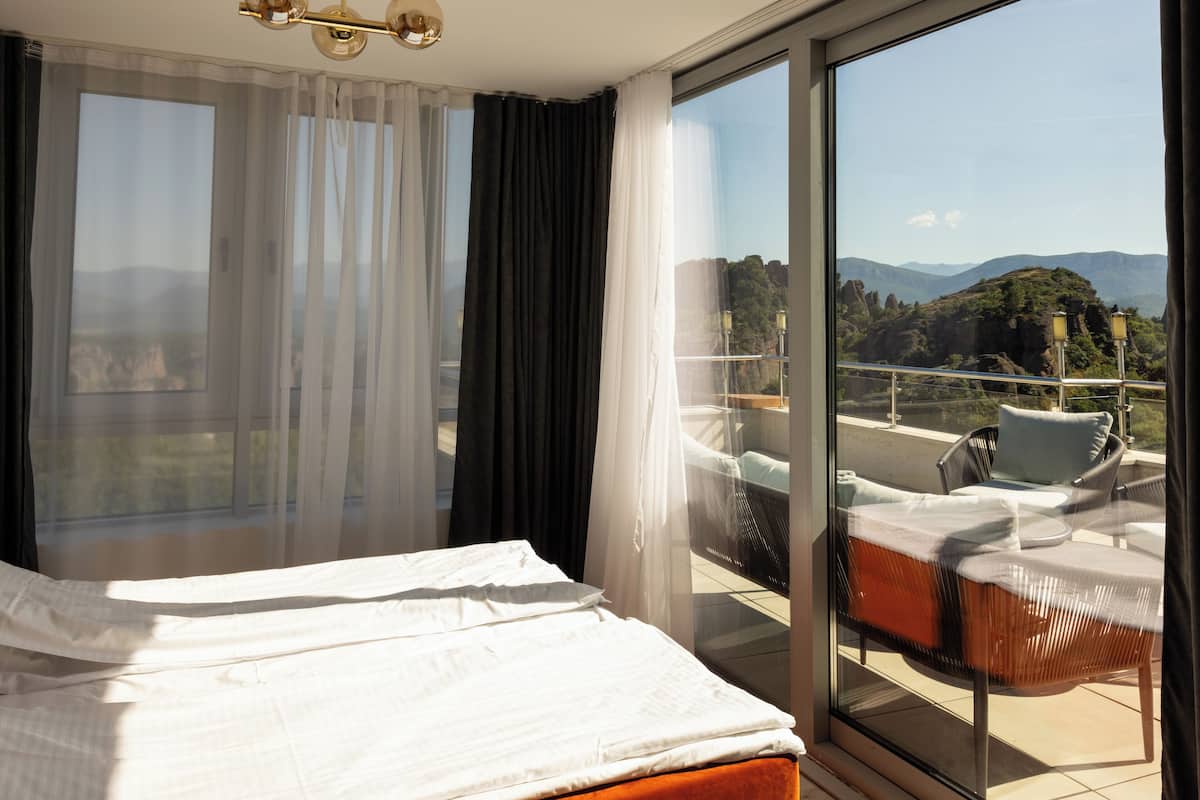 Een hotelkamer met een groot bed, een balkon met een stoel en uitzicht op de bergen.