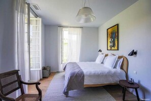 A bedroom with a bed, a chair, a small table, a painting, and a window with curtains. - Villa N1 Saint-Émilion (Saint-Émilion)