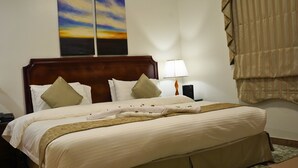 A neatly made bed with a wooden headboard, a bedside lamp, and a painting of a sunset on the wall. - Sand Rose Suite Hotel (Al Khobar)