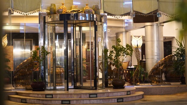 A revolving door with glass panels, surrounded by potted plants and a decorative lamp. - Sand Rose Suite Hotel (Al Khobar)