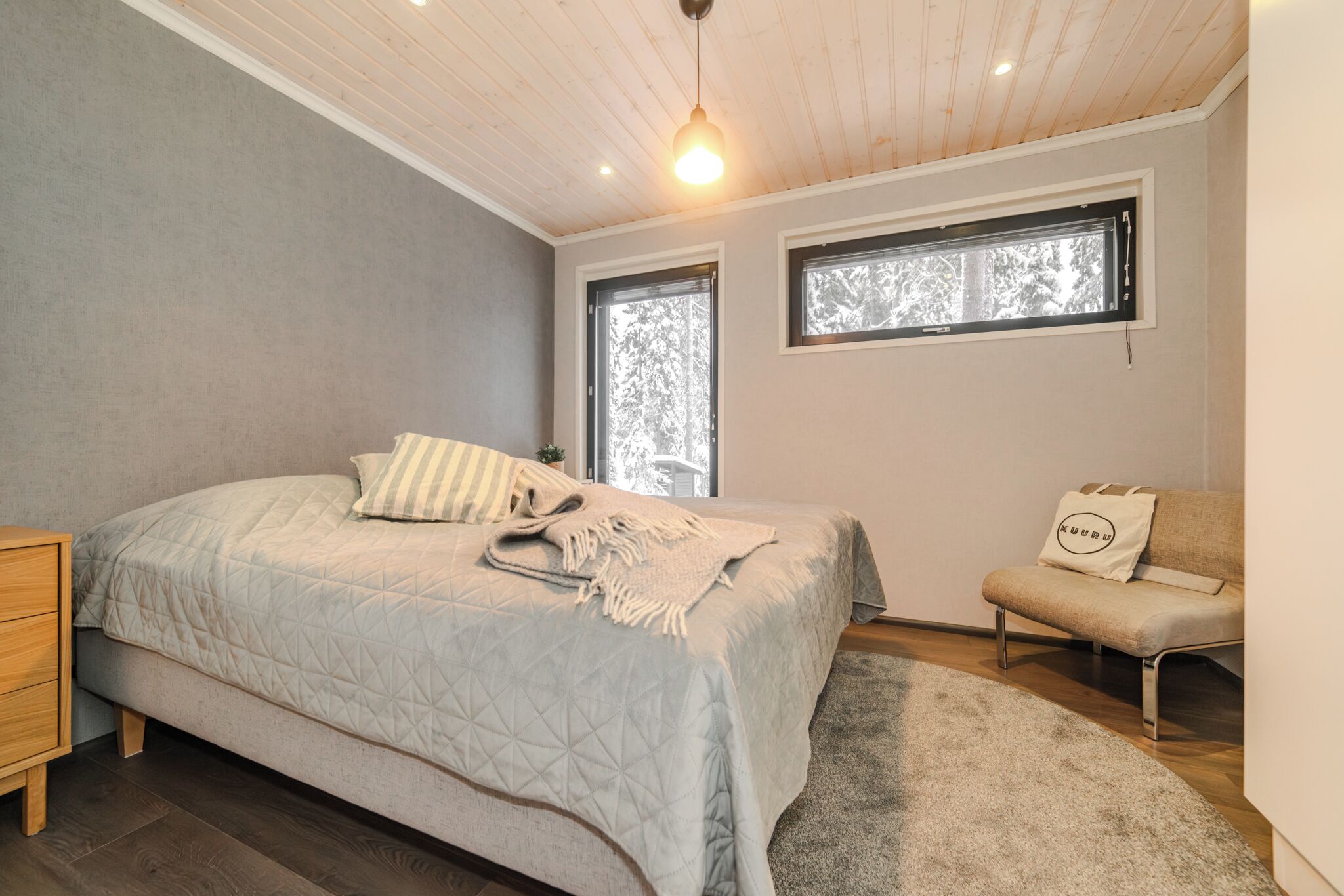 Un dormitorio con una cama, dos ventanas, techo de madera, una mesita de noche y un sofá.