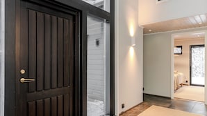 A modern interior with a dark wooden door, a glass sliding door, and a hallway leading to a bedroom with a bed and a TV. - Villa Ailo - Quality Villa with Sauna & Jacuzzi (Kuusamo)