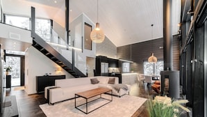A spacious living area with a large sofa, a coffee table, and a fireplace. There is a staircase leading to an upper level, and a dining area with chairs and a table. - Villa Ailo - Quality Villa with Sauna & Jacuzzi (Kuusamo)