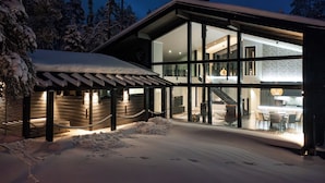 A modern house with large glass windows, a covered outdoor area, and snow on the ground. - Villa Ailo - Quality Villa with Sauna & Jacuzzi (Kuusamo)