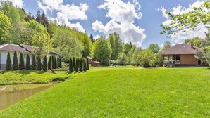 A well-maintained grassy area with a small pond, surrounded by neatly trimmed hedges and trees, and houses with sloped roofs.