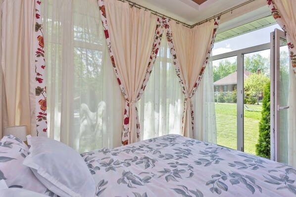 A bedroom with a large bed, floral curtains, and a view of a green landscape through a glass door.