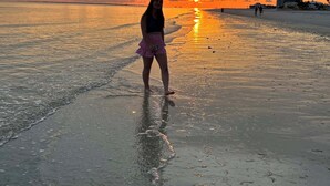 Cena de praia com um pôr do sol, uma pessoa em pé na água e edifícios ao fundo.