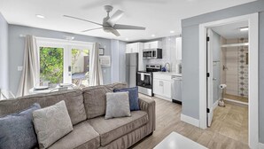 Uma sala de estar moderna com um sofá cinzento, um ventilador de teto e uma área de cozinha com armários brancos e um micro-ondas.