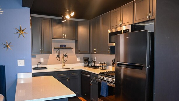 A modern kitchen with dark cabinets, a stainless steel refrigerator, a gas stove, and a white countertop.