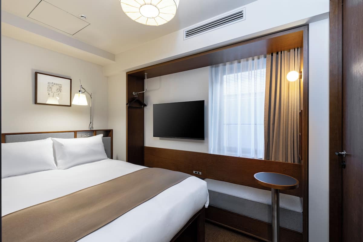 A hotel room with a bed, a TV, a bedside table, a window with curtains, and a wall-mounted picture.
