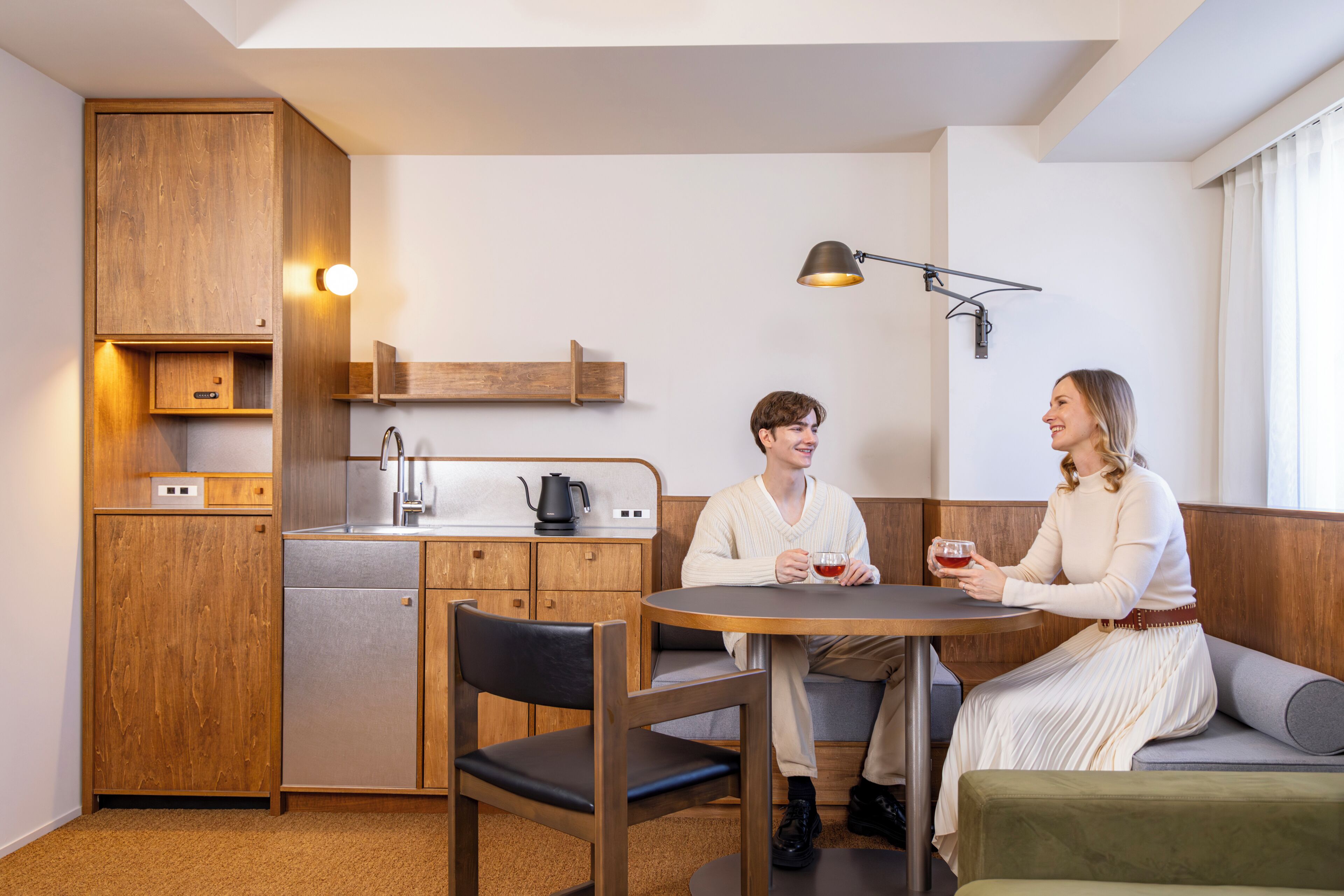 A compact kitchen with wooden cabinetry, a built-in dishwasher, and a coffee maker. There is a round table with two chairs and a bench seating area.