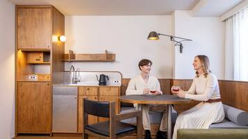 A compact kitchen with wooden cabinetry, a built-in dishwasher, and a coffee maker. There is a round table with two chairs and a bench seating area.