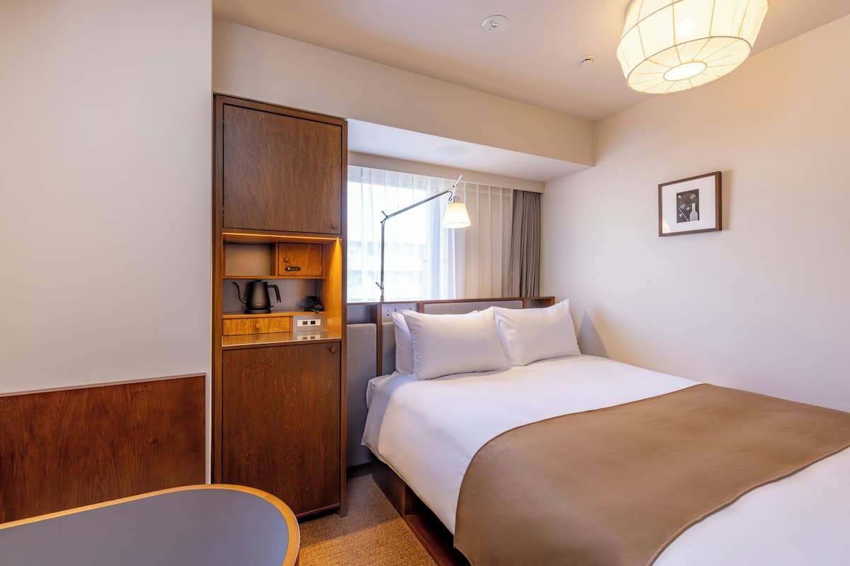 A hotel room with a bed, a wooden wardrobe, a desk, a lamp, and a window with curtains.