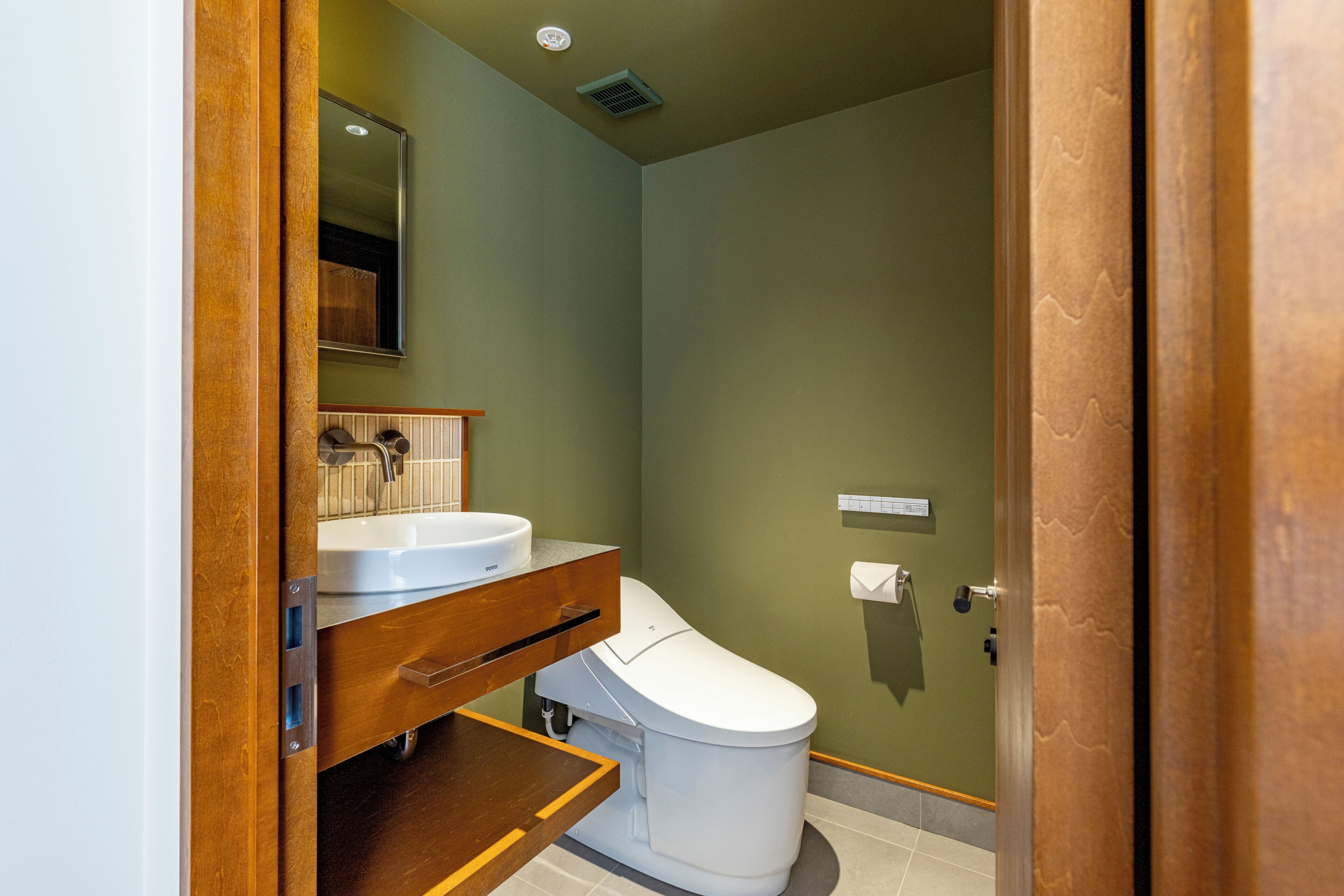 A bathroom with a white toilet, a wooden vanity, a mirror, and a wall-mounted toilet paper holder.